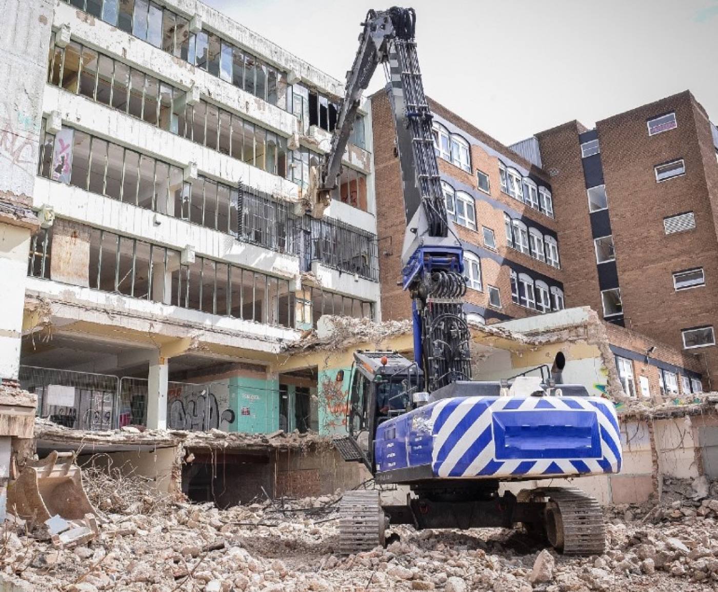 A high-reach demolition machine working on a multi-storey concrete frame, its
                 façade and floor slabs partly removed, with the arisings graded below.